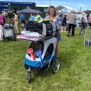 Medium Petstro dogshow stroller in blue, with grooming table top fully loaded & being pushed by a lady at a dog show